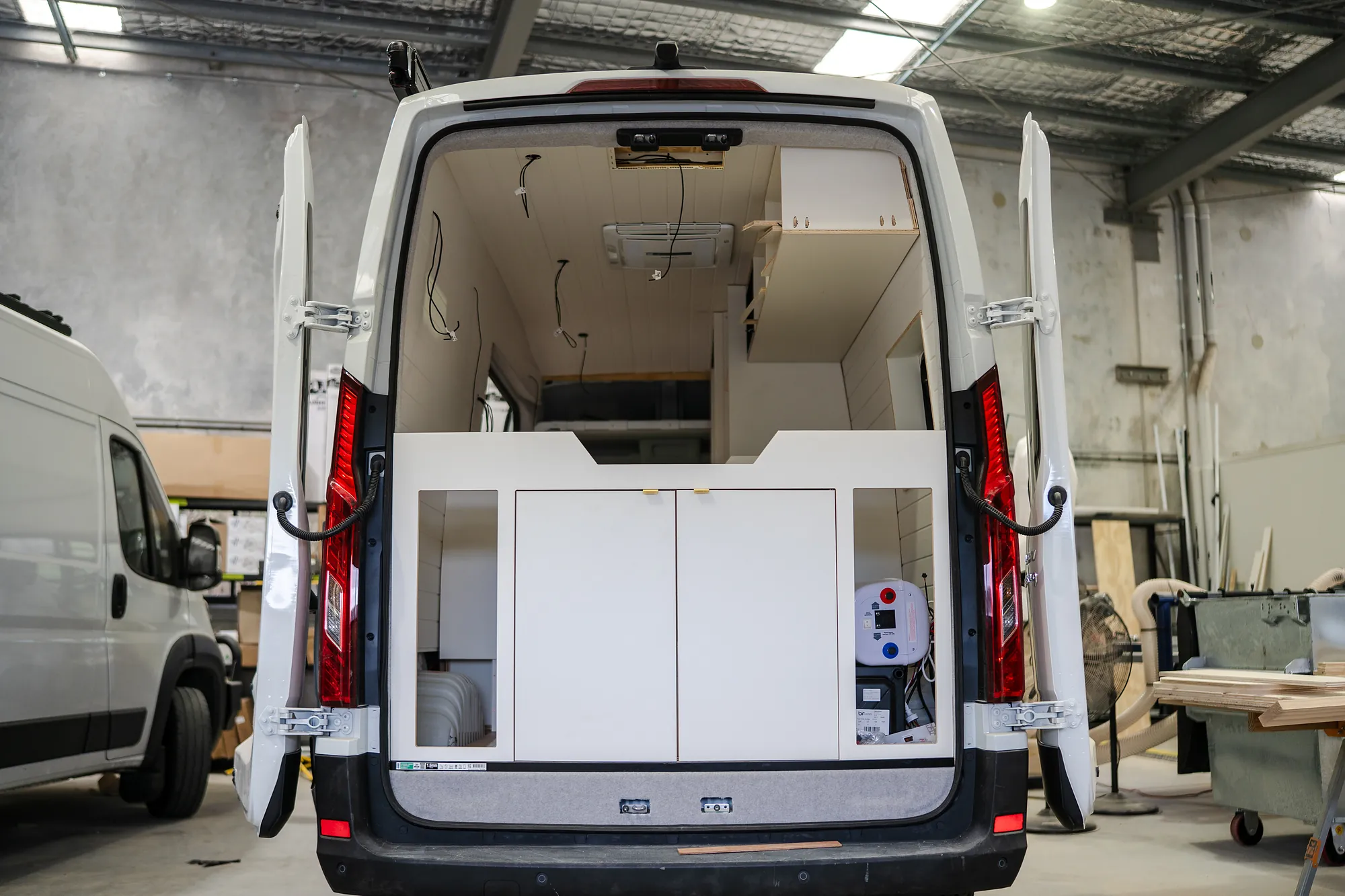 High-roof campervan with rear doors open in the workshop, CNC plywood rear cabinet and overhead storage fitted