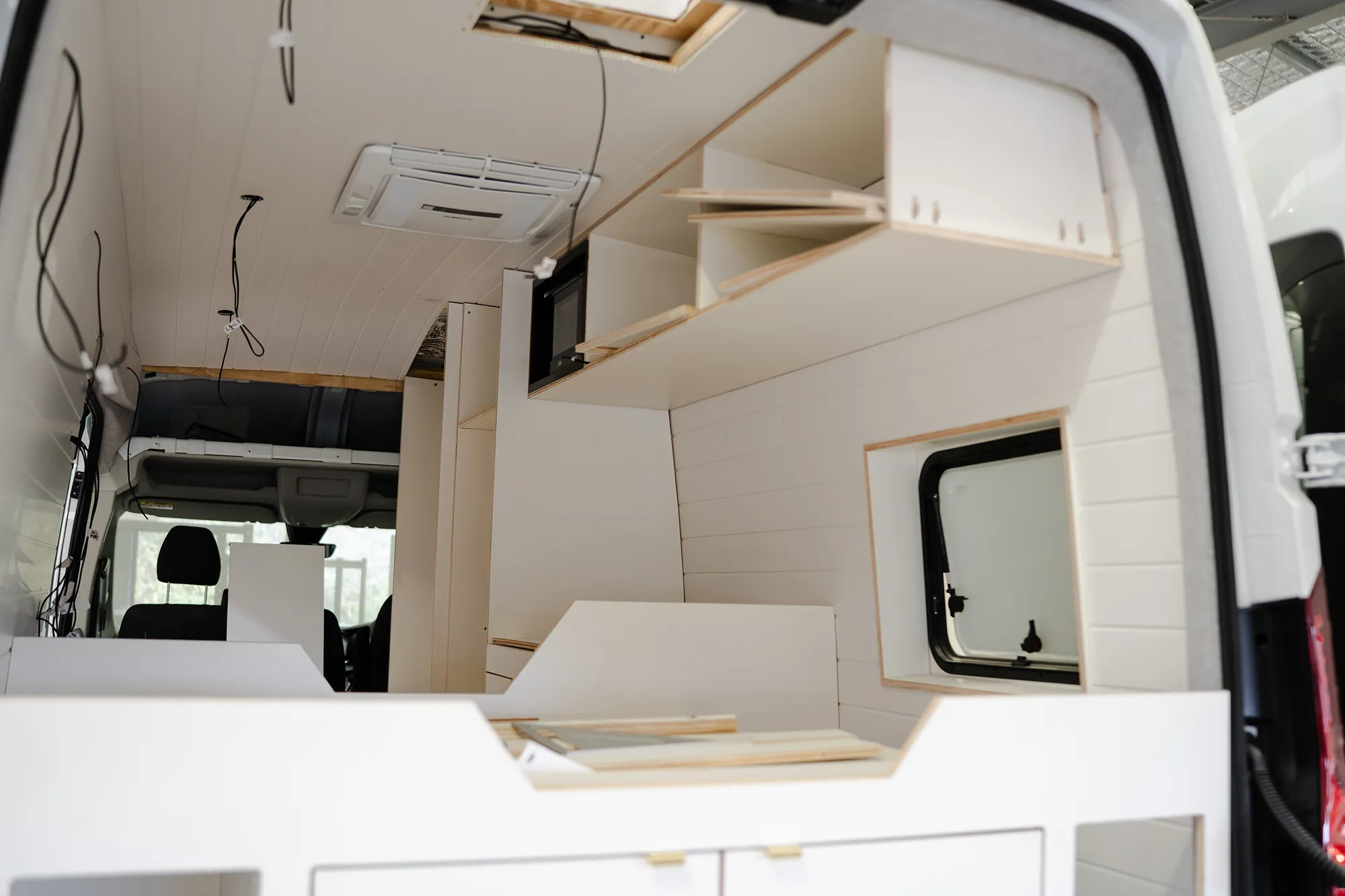 Wide view of the campervan interior from the rear doors: overhead storage, kitchen unit and bed platform in white plywood